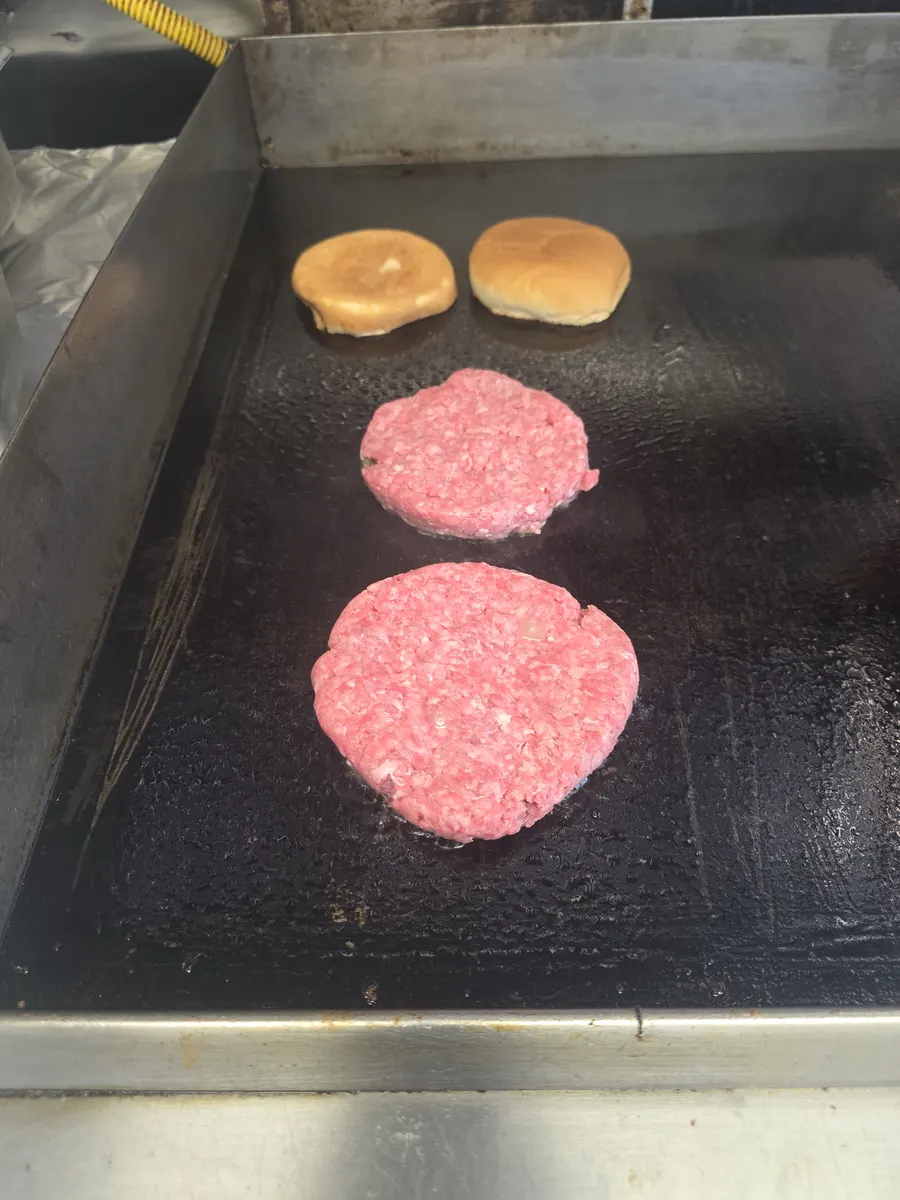 Two fresh Angus patties on the flat top with buns toasting behind them