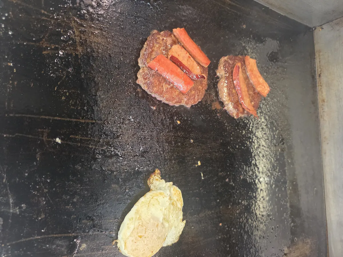 Both patties topped with hotlink while a fried egg cooks beside them