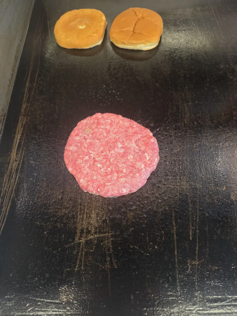 A fresh beef patty and two bun halves on the flat top