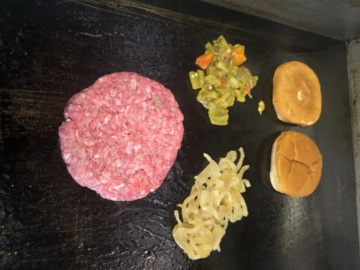 The patty on the griddle beside chopped Hatch green chiles, grilled onions and the buns