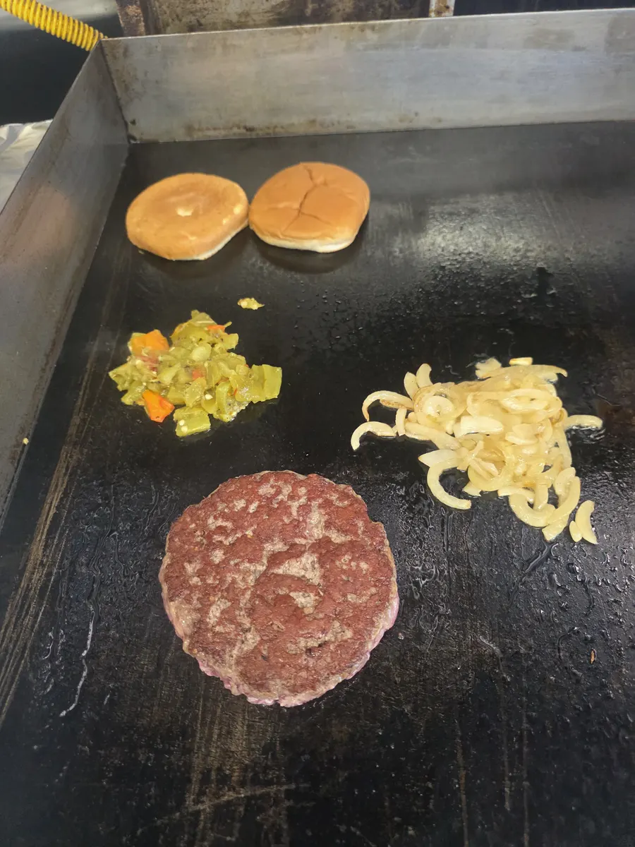 The patty seared and flipped, green chile and grilled onions working beside it
